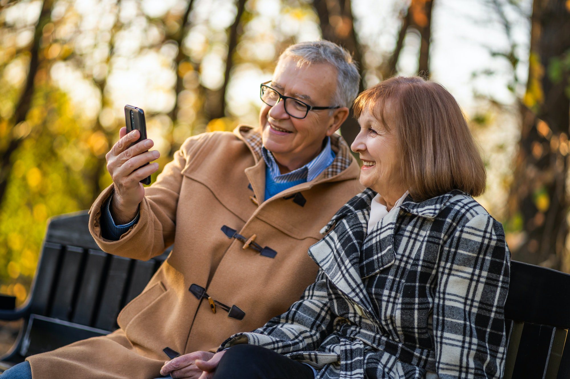 Happy senior couple enjoying fall weather sitting on park bench taking a picture and discussing when their medicare will start.